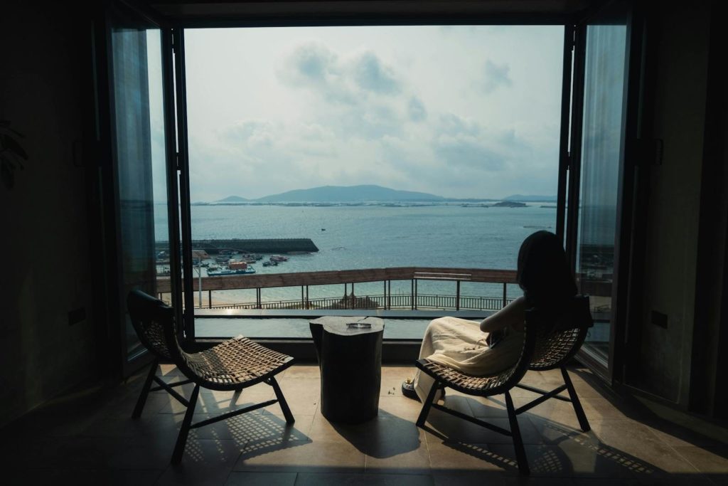 A woman sits by a large open window enjoying a serene ocean view, surrounded by modern wicker furniture.